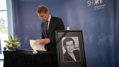 Ministerpräsident Günther blättert durch ein Kondolenzbuch. Im Vordergrund steht das Bild von Heide Simonis auf einem dunkel geschmückten Tisch neben einer Kerze.