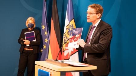 Ministerpräsident Daniel Günther steht an einem Rednerpult. Im Hintergrund steht Michaela Pries, die Landesbeauftragte für Menschen mit Behinderung. 