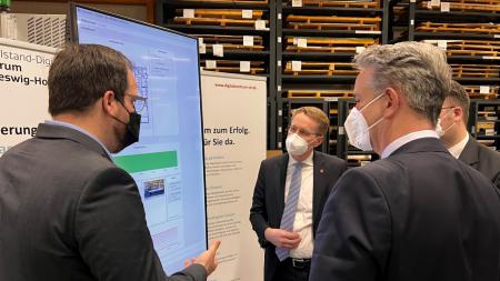 Ministerpräsident Daniel Günther und mehrere Angestellte der Walter Otto Müller GmbH schauen auf einen Bildschirm.