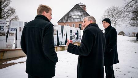 Ministerpräsident Daniel Günther, der Historiker Uwe Danker und Bischof Gothart Magaard stehen gemeinsam auf dem Außengelände des Lernorts und betrachten die Ausstellung.