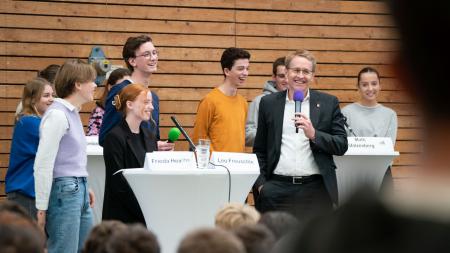 Ministerpräsident steht vor einer Gruppe von Schülerinnen und Schülern und hält ein Mikrofon in der Hand.