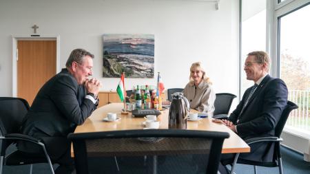 Ministerpräsident Daniel Günther sitzt dem ungarischen Botschafter Dr. Peter Györkös während eines Gesprächs an einem Tisch gegenüber. Eine Frau verfolgt im Hintergrund das Gespräch.