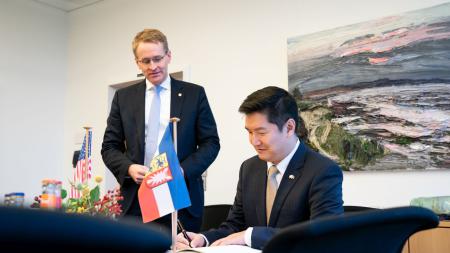 Der Generalkonsul der USA Jason Chue sitzt an einem Tisch und trägt sich in ein Buch ein. Ministerpräsident Daniel Günther steht links daneben.