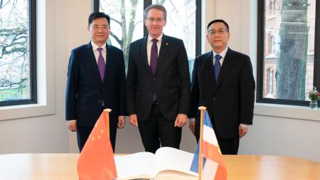 Ministerpräsident Daniel Günther steht zwischen zwei Männern. Auf dem Tisch vor der Gruppe liegt ein aufgeschlagenes Buch und die Flaggen der Volksrepublik China und Schleswig-Holsteins.