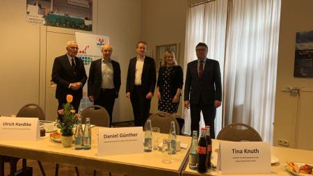 Ministerpräsident Daniel Günther (3.v.l.) nimmt gemeinsam mit vier weiteren Personen Aufstellung für ein Gruppenbild. Im Vordergrund ist ein Tisch mit Namensschildern zu sehen.