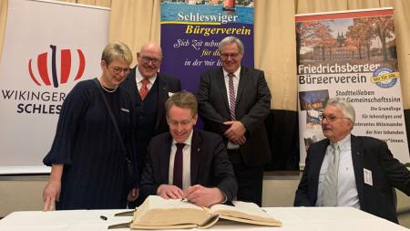 Ministerpräsident Daniel Günther sitzt an einem Tisch und schreibt in ein großes Buch. Um ihn herum stehen drei Vertreterinnen und Vertreter der Stadt Schleswig, darunter auch Bürgermeister Dose.
