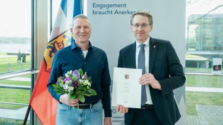 Ministerpräsident Daniel Günther und Andreas Euker stehen nebeneinander. Der Ministerpräsident hält die Urkunde in den Händen und Andreas Euker einen Blumenstrauß.