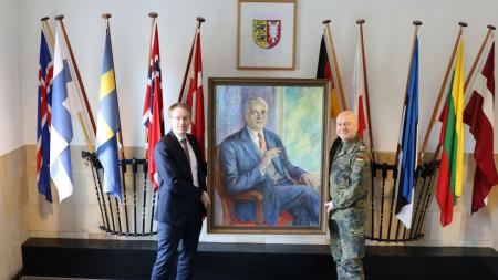 Ministerpräsident Daniel Günther und Oberst Axel Schneider präsentieren ein Porträt Theodor Steltzers. Im Hintergrund hängt das Wappen des Landeskommandos Schleswig-Holsteins sowie Flaggen zahlreicher europäischer Staaten.