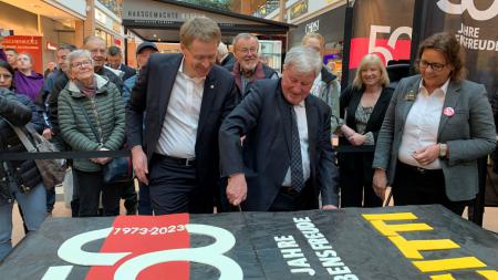 Ministerpräsident Daniel Günther schneidet gemeinsam mit einem Mann einen großen Kuchen an. Im Hintergrund stehen zahlreiche Schaulustige.