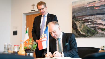 Ministerpräsident Daniel Günther steht hinter einem Mann, der sich an einem Tisch sitzend in ein Buch einträgt. Auf dem Tisch stehen die Flaggen Schleswig-Holsteins und Irlands.