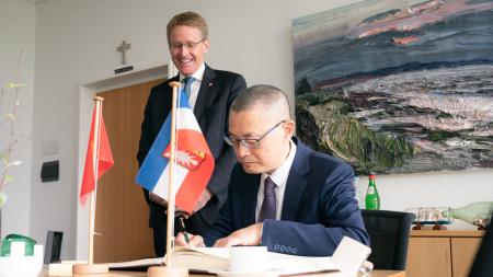 Ministerpräsident Daniel Günther steht hinter dem vietnamesischen Botschafter, der sich an einem Tisch sitzend in das Gästebuch einträgt.
