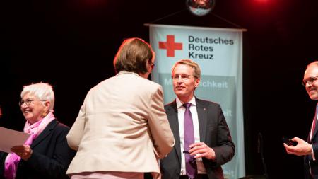 Ministerpräsident Daniel Günther gibt einer Frau die Hand. Links daneben steht eine weitere Frau, die eine Urkunde hält.
