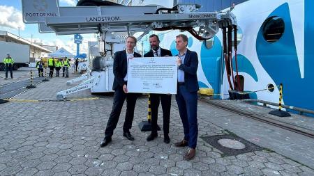 Daniel Günther, Dirk Clause und Ulf Kämpfer stehen vor einem Kreuzfahrtschiff, das an eine Landstromanlage angeschlossen ist. Sie halten gemeinsam ein Schild in die Höhe, das auf die Förderung des Landes hinweist.