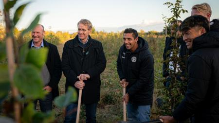 Ministerpräsident Daniel Günther steht mit anderen Männern in einem Waldstück. Er stützt sich auf einen Spaten und lächelt in die Kamera.