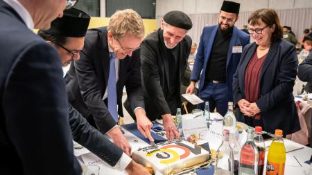 Ministerpräsident Daniel Günther schneidet einen Kuchen an, mehrere Männer stehen um ihn herum. Von der Seite schaut Vize-Landtagspräsidentin Eka von Kalben dabei zu.