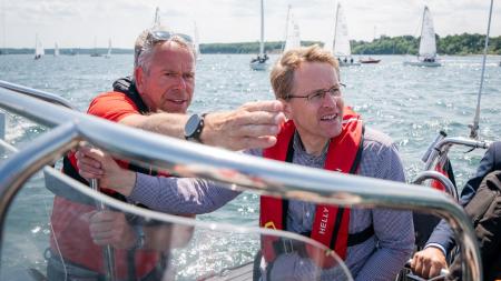 Regatta-Chef Dirk Ramhorst und Ministerpräsident Daniel an Bord eines Bootes in der Kieler Förde. Ramhorst zeigt dem Ministerpräsidenten etwas und zeigt darauf. Im Hintergrund sind zahlreiche Segel zu erkennen.