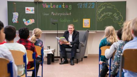 Ein Mann sitzt in einem Klassenraum und liest Kindern aus einem Buch vor.
