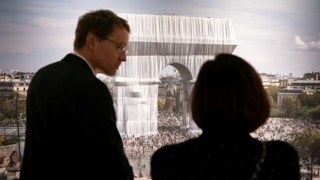 Ministerpräsident Daniel Günther und eine Frau stehen vor einer mannshohen Fotografie des verhüllten Triumphbogens in Paris.
