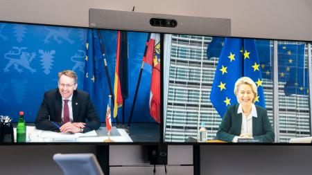 Ministerpräsident Daniel Günther steht vor einem Bildschirm und schaut in die Kamera. Auf dem Bildschirm ist EU-Kommissionspräsidentin Ursula von der Leyen zu sehen.