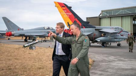 Ministerpräsident Günther läuft mit einem Soldaten über ein Flugfeld. Im Hintergrund stehen Kampfflugzeuge mit der Aufschrift Air Defender 2023