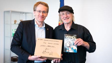 Ministerpräsident Daniel Günther und der Comiczeichner Rötger Feldmann stehen nebeneinander im Amtszimmer des Ministerpräsidenten. Sie halten ein Exemplar des berühmten Werner-Comics in die Kamera.