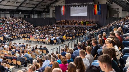 Blick in die voll besetzte Holstenhalle in Neumünster. Auf einer Bühne steht Ministerpräsident Daniel Günther und hält eine Rede. Im Publikum sitzen angehende Polizeibeamte sowie deren Angehörigen. 
