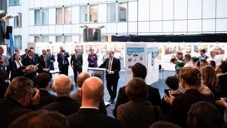 Ministerpräsident Daniel Günther steht an einem Rednerpult inmitten einer Ausstellung. Um ihn herum stehen zahlreiche Menschen und lauschen seiner Rede.