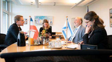 Ministerpräsident Daniel Günther und der israelische Botschafter Professor Ron Prosor sitzen gemeinsam mit zwei Frauen an einem Tisch. Auf diesem stehen die Flaggen Israels und Schleswig-Holsteins.