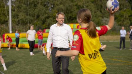 Ministerpräsident Daniel Günther steht auf einem Handball-Feld. Vor ihm steht eine Spielerin und wirft ihm einen Ball zu. 