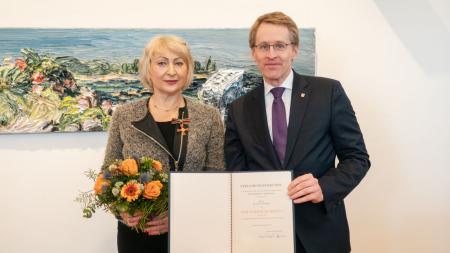 Ministerpräsident Daniel Günther und die Ausgezeichnete stehen in einem Raum vor einem großen Gemälde. Die Geehrte trägt Orden an der Kleidung und einen Blumenstrauß in den Händen. Der Ministerpräsident hält ihre Urkunde in den Händen.