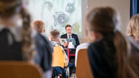 Ministerpräsident Daniel Günther sitzt vor einer Schulklasse und liest den Kindern aus einem Buch vor.