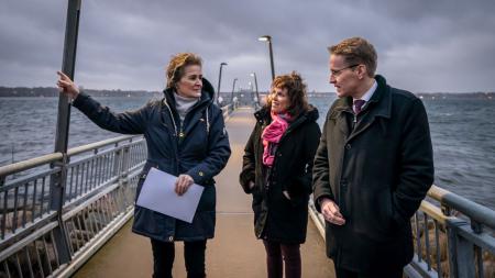 Ministerpräsident Daniel Günther, Plöns Landrätin Stephanie Ladwig und Mönkebergs Bürgermeisterin Hildegard Mersmann stehen an einem Fähranleger.
