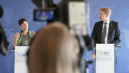 Finanzministerin Heinold und Ministerpräsident Günther stehen an Rednerpulten und werden dabei von einer Kamera gefilmt.