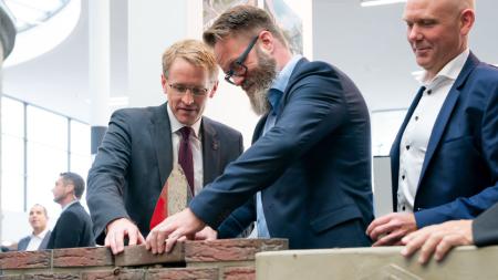 Ministerpräsident Günther und Wirtschaftsminister Madsen passen einen Ziegelstein in eine Mauer ein. 