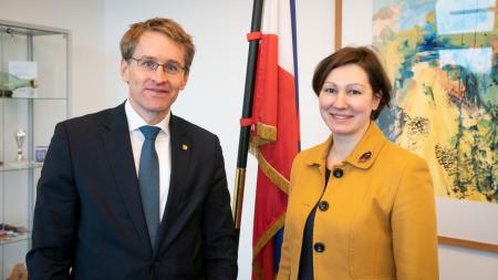 Daniel Günther und die lettische Botschafterin stehen nebeneinander. Im Hintergrund ist die Flagge Schleswig-Holsteins zu sehen. 