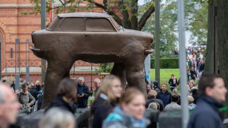 Eine große Statue eines Autos mit vier Beinen steht auf einer Grünfläche, darum herum laufen Menschen. 
