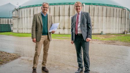 Ministerpräsident Daniel Günther und der Marienthal-Geschäftsführer Philip Klagges stehen nebeneinander. Klagges hält eine Mappe in den Händen, hinter den beiden ragt eine Biogasanlage in den Himmel.